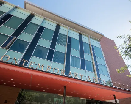 A low-angle view of the Contemporary Arts Center, with bronze letters spelling its name along a red-orange entrance canopy, a facade of teal and navy glass panels and a red brick wall against a clear blue sky.