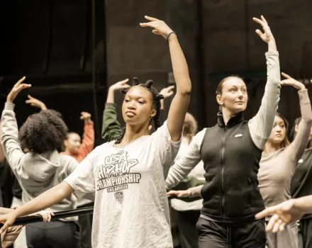 Dance students rehearse choreography in a studio, extending their arms in coordinated poses.