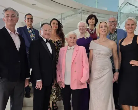 Nine OC Arts Awards honorees in formal attire pose together on a staircase with glass railings in a bright, modern lobby. A grand piano is visible behind them.