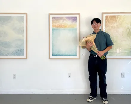 An artist holding a bouquet of flowers smiles beside three framed works on a white gallery wall. The works are soft, layered abstractions in blue, gold and green that suggest water and sky.