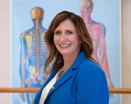 Dr. Kelli Sharp smiles at the camera, wearing a bright blue blazer over a white blouse. Anatomical illustrations of the human skeletal and muscular systems are displayed on the wall behind her.