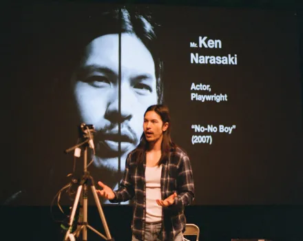 Kurt Kana, wearing a plaid shirt over a white tank top, speaks on stage with his hands open beside a camera on a tripod. A projection behind him shows a black-and-white close-up of a face and the words "Mr. Ken Narasaki, Actor, Playwright, 'No-No Boy' (2007)."