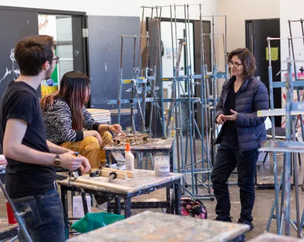 Professor Monica Majoli talks with two students in a paint-spattered studio filled with metal easels. One student sits at a worktable assembling a small brass and metal sculpture, while the other stands nearby holding a tool.