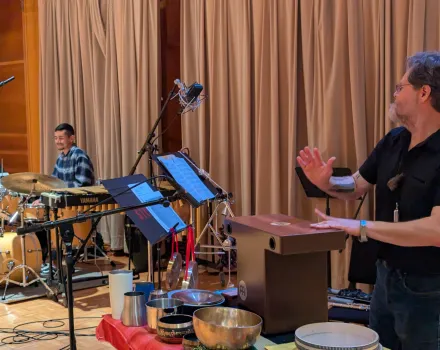 Camilo Zamudio plays marimba behind a drum kit as co-leader Wilfrido Terrazas gestures beside a cajón and a table of singing bowls and frame drums during a rehearsal in a wood-paneled studio.