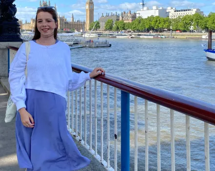 Emily Parise, in a white sweater and periwinkle dress, smiles beside a railing on the River Thames, with Big Ben and the Houses of Parliament in the background.