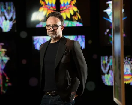 Matt Bailey, wearing a dark blazer over a black T-shirt and black-framed glasses, stands with one hand in his pocket in a darkened gallery surrounded by glowing, symmetrical digital artworks in vivid rainbow colors.
