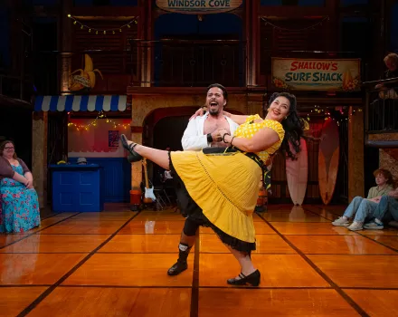 On a sunlit wooden stage dressed as a beachside boardwalk, with signs for “Windsor Cove” and “Shallow’s Surf Shack,” a laughing man in an open white shirt dips a smiling woman in a yellow polka-dot top and full yellow skirt as she kicks one leg high. Audience members seated around the stage edge look on and smile.