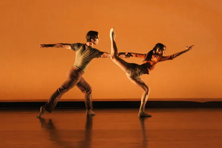 Two dancers perform on a stage washed in warm orange light, moving in a dynamic partnered pose. One dancer extends forward while the other lifts a leg high behind them, both reaching outward to emphasize balance, tension, and fluid motion.