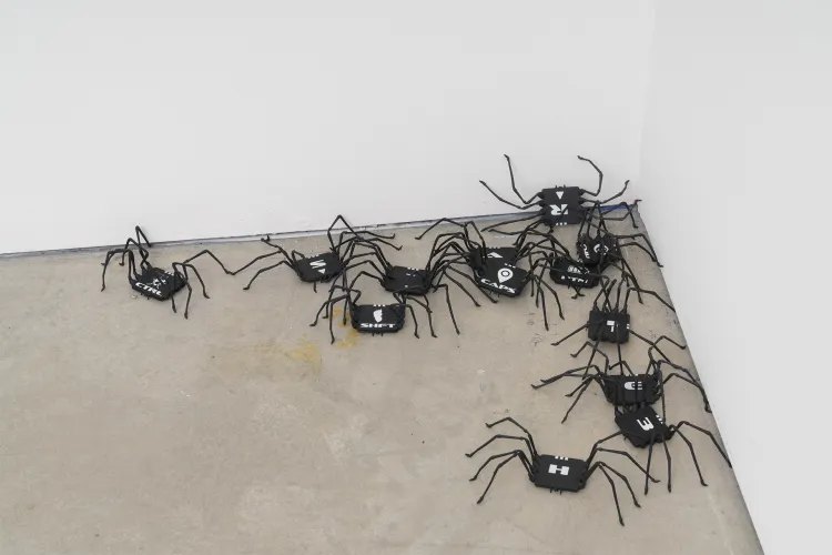 Cluster of small black spider-like sculptural objects arranged along the corner of a gallery floor and wall, each with thin wire legs and box-like bodies.