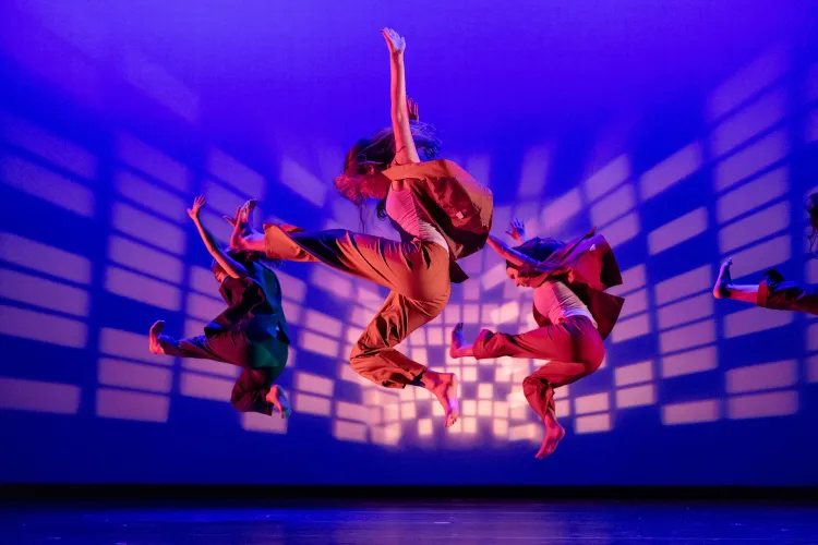 Group of dancers mid-jump on a stage under colorful lighting, captured in synchronized airborne movement against a patterned light backdrop.
