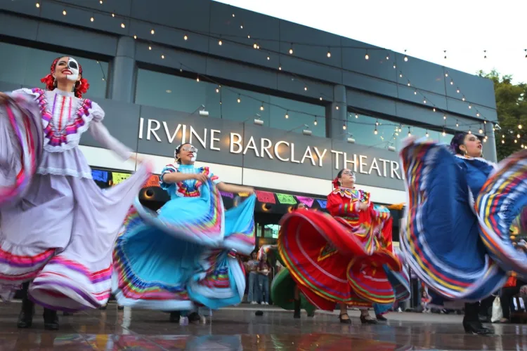 Dancers in vibrant, traditional folklórico dresses perform outside the Irvine Barclay Theatre. Their colorful skirts flare outward as they spin beneath string lights, with the theater’s name visible on the building behind them.