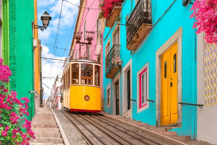 A bright yellow tram travels up a steep cobblestone street lined with colorful buildings, balconies, and pink flowers, under a clear blue sky.