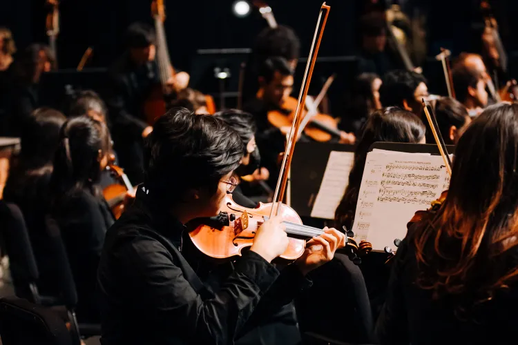 Orchestra musicians seated on stage play violins while reading sheet music under warm stage lighting.