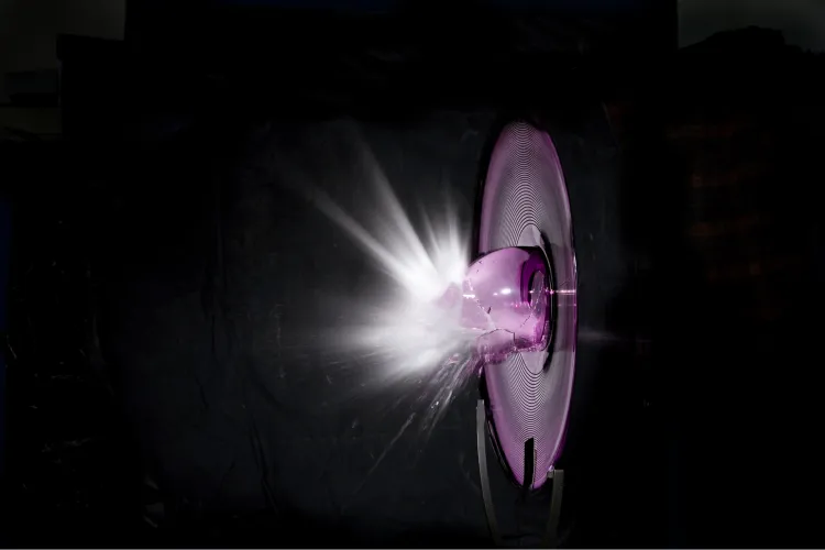 A bright beam of light shines onto a circular glass or metal disc in a dark setting. The disc reflects the light in a radiant burst, creating a glowing focal point against the black background.