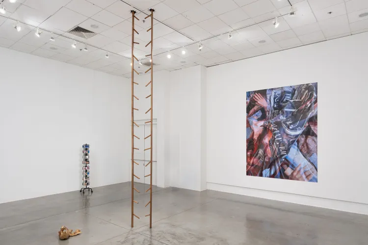 A contemporary gallery installation featuring a tall ladder-like sculpture and a large mixed-media painting displayed in a white exhibition space.
