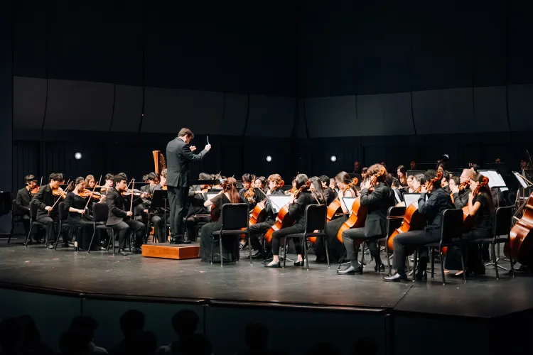 A full orchestra performs on a concert hall stage, with a conductor standing on a podium leading the musicians. String players fill the foreground and sides, seated with music stands, while additional sections extend across the stage, all dressed in formal black under focused stage lighting.