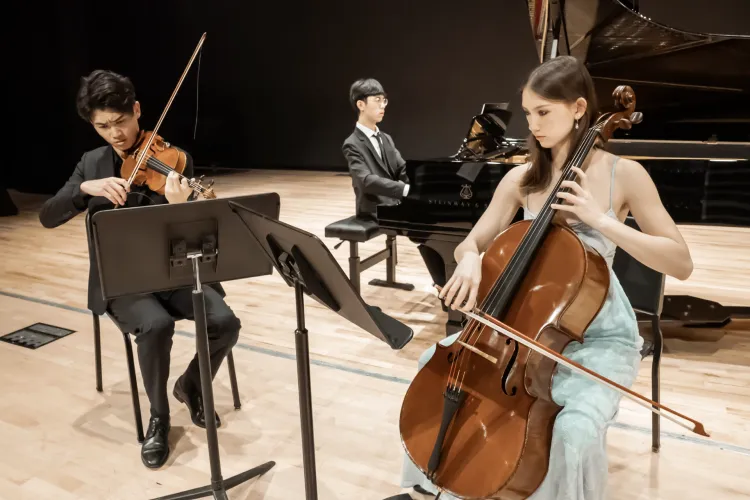 Three musicians perform on a concert stage, with a violinist and cellist reading from music stands and a pianist seated at a grand piano behind them.