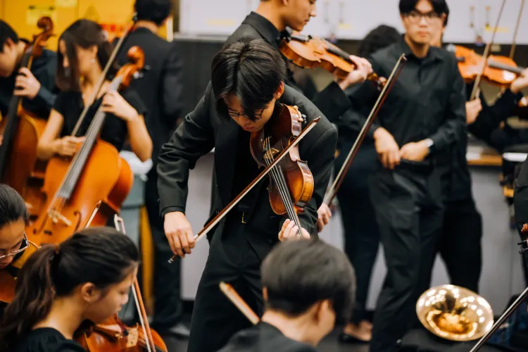 A violinist in black formal attire leans forward while playing, surrounded by other string and brass musicians performing together in an orchestra ensemble.