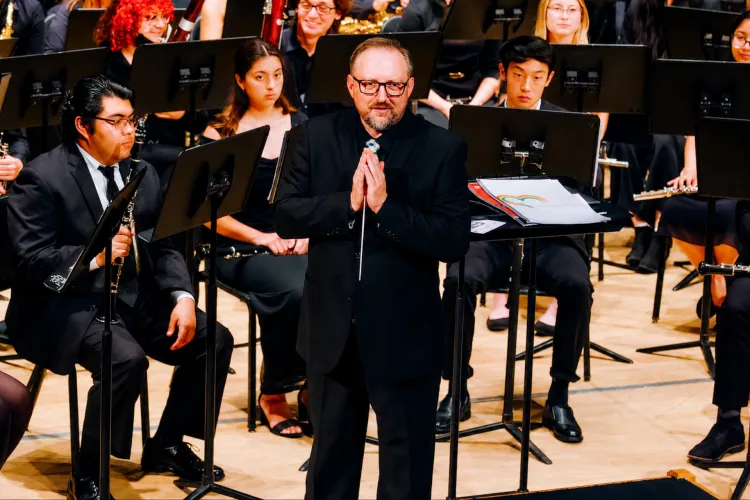 Kevin McKeown in black formal attire holds a baton with hands clasped while standing before a seated ensemble of student musicians with music stands on a concert stage.