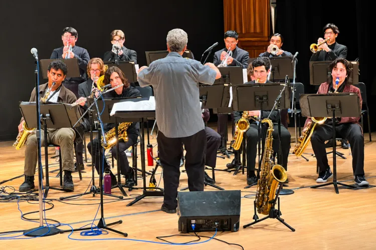 A conductor leads a jazz ensemble of approximately a dozen student musicians playing saxophones, trumpets and trombones on a wood-floored stage with music stands and microphones.