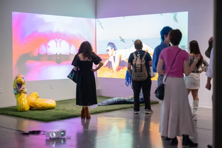 Gallery visitors stand before large-scale video projections spanning two walls, featuring close-up lips and a beach scene, alongside a sculptural yellow snake on artificial turf.