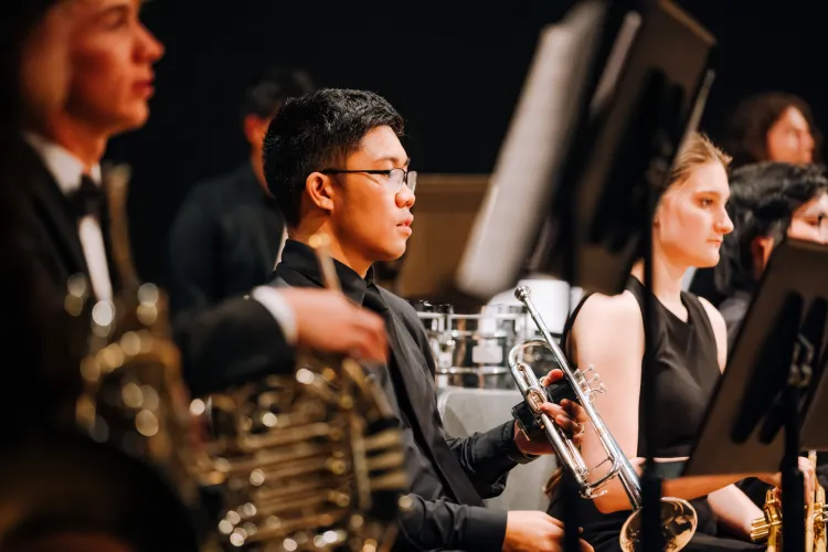 A musician wearing glasses holds a silver trumpet while seated among other formally dressed ensemble members with brass instruments and music stands.