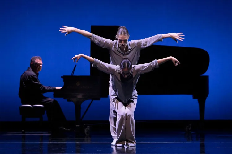 Two dancers perform synchronized movements in front of a pianist and grand piano under blue stage lighting.