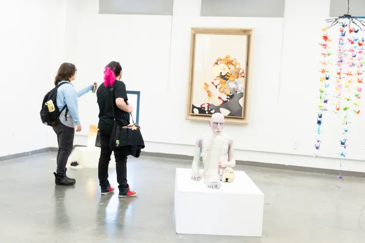 Two gallery visitors view artwork in a white exhibition space featuring a figurative sculpture on a pedestal, a framed painting, and a hanging colorful origami crane installation.