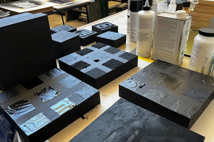 Multiple black relief printing blocks with textured, inked surfaces arranged on a worktable in a printmaking studio, with bottles of printing medium nearby.