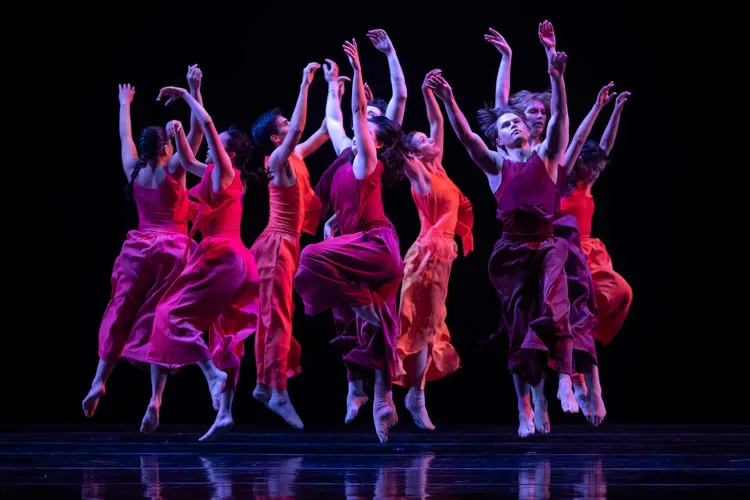 A group of dancers leap and move together on stage in flowing red costumes during a contemporary dance performance.