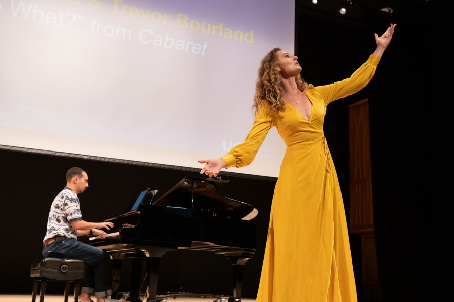 Woman in a yellow dress singing with her arms extended and accompanied by a man at the piano