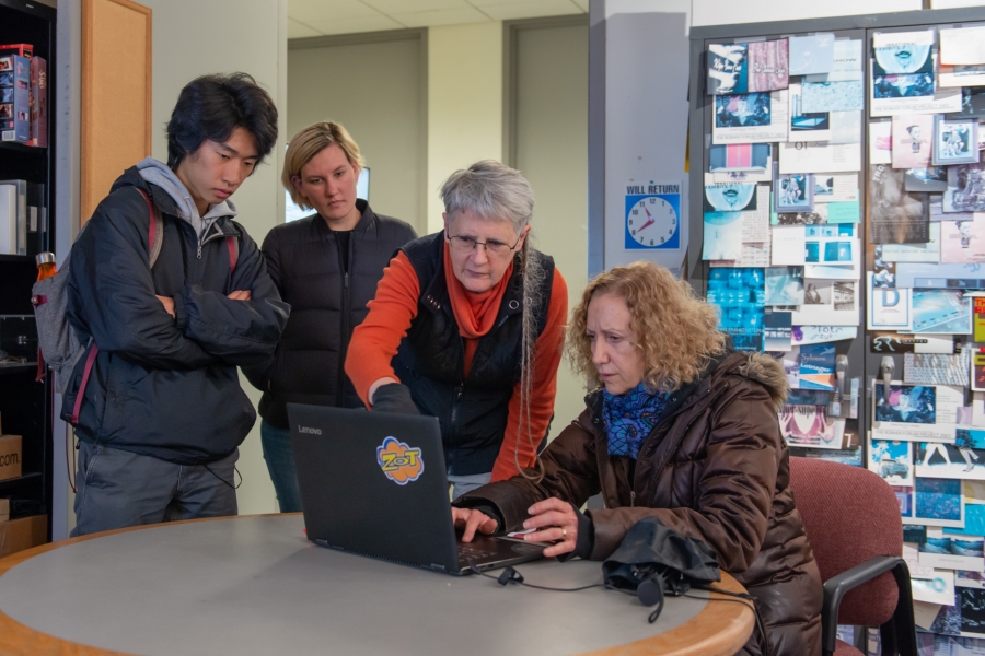 A professor sits down at a table with an older professor standing above them pointing at a computer