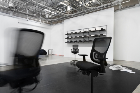 A gallery installation with office chairs, shelving, papers on the floor