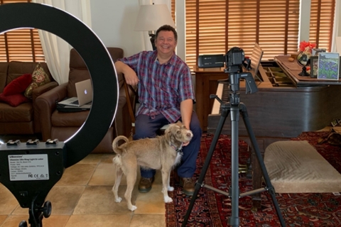 Alan Terricciano sitting at piano with a dog at his feet; lighting and filming equipment can be seen