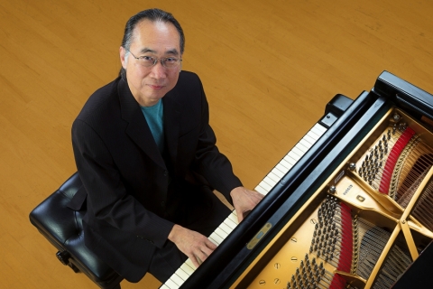 Kei Akagi sitting at piano, hand on keyboard, smiling into camera