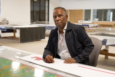 Black man seated behind what appears to be a drafting table; large-scale art-prints, or perhaps maps, displayed