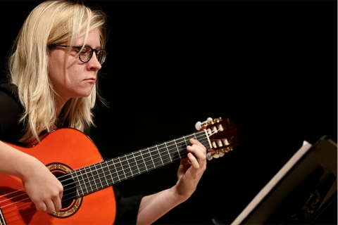 Blonde person with glasses, sittging down, playing guitar