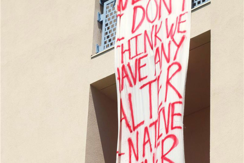 A cloth banner with large words in red hang over the side of a building balcony. 