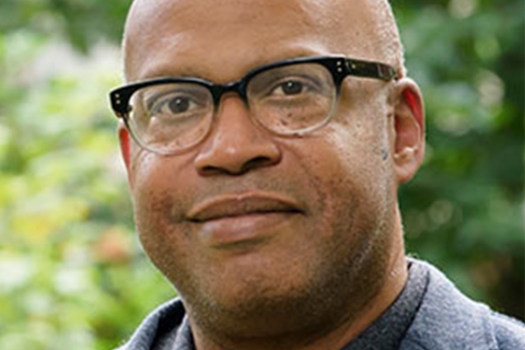 Closeup image of Black man in glasses, smiling, wearing a suit; outdoor greenery in the background