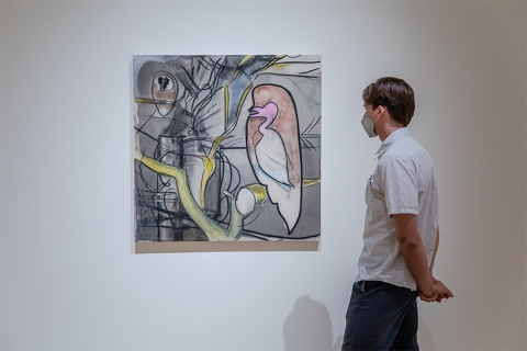 Man standing in gallery looking at painting, mask on, hands behind back