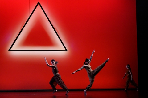 Image of dancers on stage with red background, a triangle seemingly floating mid-air