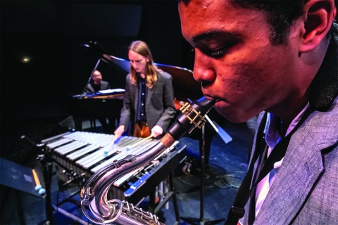 Close up of someone playing saxophone, with percussion and piano in the background