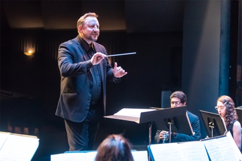 Image of music conductor with baton in hand, gesturing to students with music stands in front of them holding sheet music