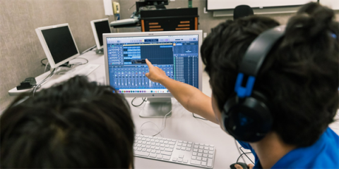 Computer screen with audio editing software controls, a student with headphones pointing to monitor