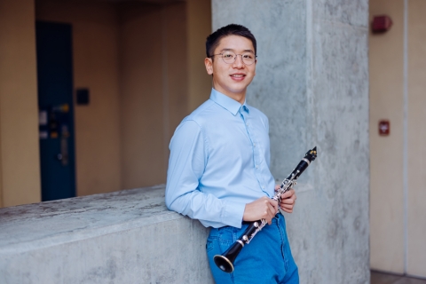 Young man with glasses holding clarinet, leaning against a concrete wall
