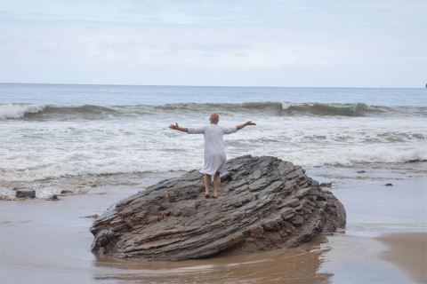 Image of bald person in tunic-like garment, standing with arms outstretched, back to camera, on a large rock on a beach with crashing waves